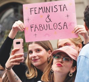 en la fotografía dos mujeres, una sostiene un letrero que dice "feminista y orgullos" y otra se toma una selfie