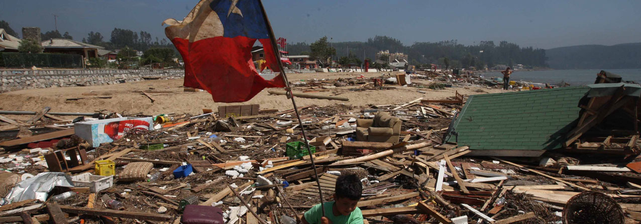 niño sostiene una bandera chilena dota y en el fondo ruinas de casas derrumbadas por un terremoto